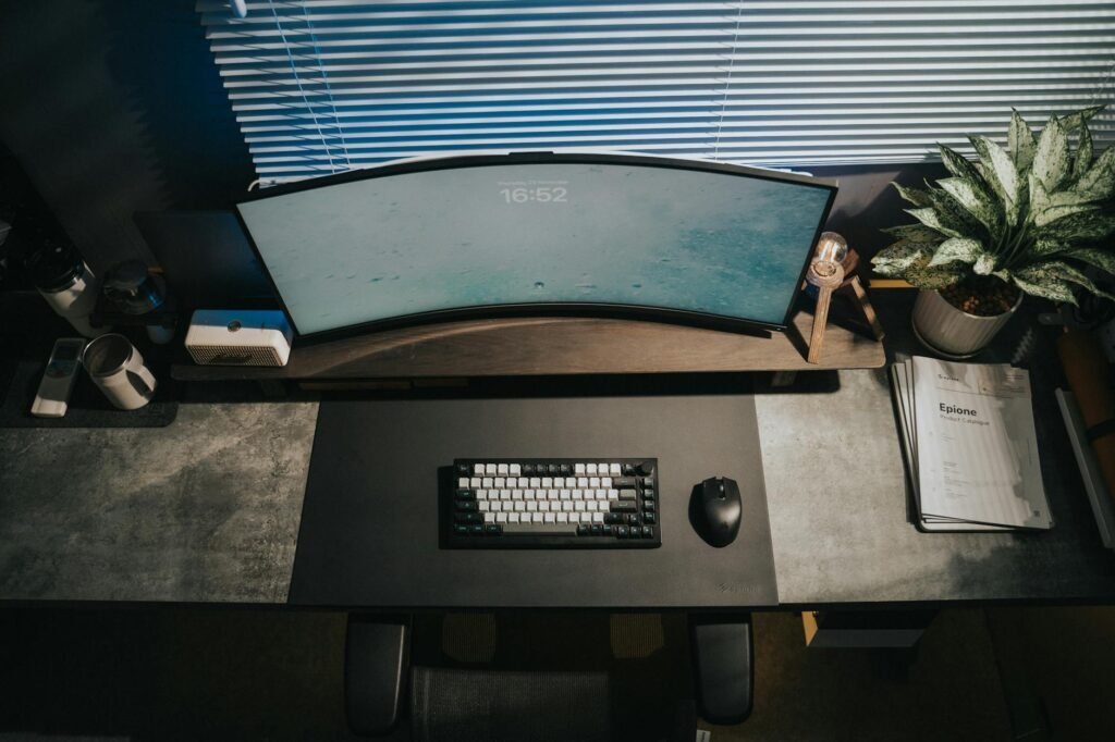 A compact desk with a monitor riser, keyboard, mouse, and neatly stored small accessories underneath the screen.